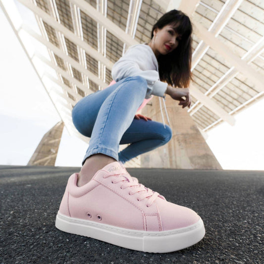 Person crouching outdoors on pavement wearing light pink Fuego low-top sneakers with white soles, photographed from a low angle to highlight the shoe against an urban architectural backdrop.