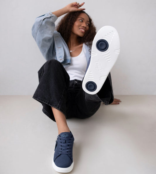 Person sitting casually against a light background wearing navy Fuego low-top sneakers, lifting one foot to show the white sole with two circular pivot points.