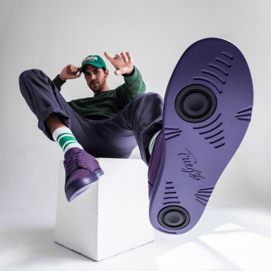 Person sitting on a white cube wearing purple Fuego low-top sneakers lifts one foot to show the sole with two circular pivot points, with a matching casual outfit and green cap against a light background.