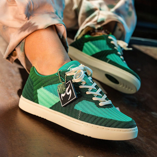 Person wearing jade green Fuego low-top sneakers with white laces and soles, with a branded tag hanging from the shoe, resting casually on a surface in warm lighting.